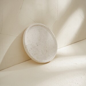 May include: A circular, off-white stone tray with a smooth, polished surface and natural veining patterns. The tray features a raised rim and a flat base, showcasing a consistent light beige color palette with subtle, organic mineral textures.