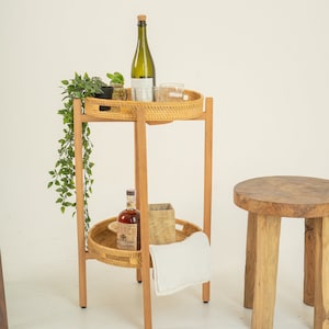 Könnte beinhalten: Ein zweistöckiger Servierwagen aus Holz mit einer geflochtenen Rattan-Oberfläche und einem unteren Regal. Der Wagen hat eine Flasche Wein, eine Flasche Spirituosen, ein Glas und eine Topfpflanze auf der oberen Ablage. Ein weißes Handtuch und eine braune Papiertüte befinden sich auf der unteren Ablage. Ein Holzhocker steht im Hintergrund.
