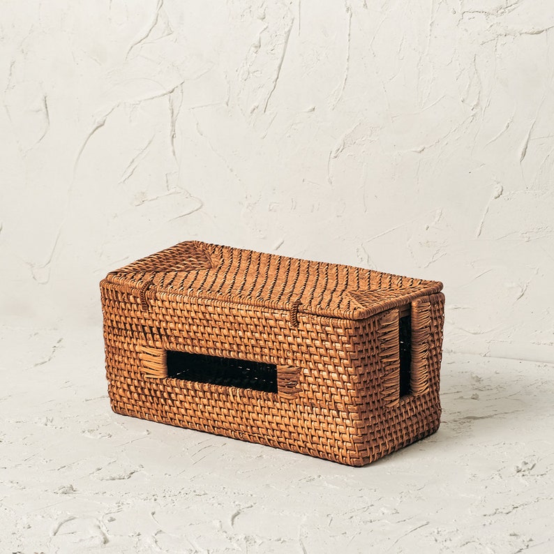 May include: A woven wicker tissue box cover with a rectangular shape and a cut-out opening for tissues. The cover is made of natural brown wicker and has a slightly textured surface.