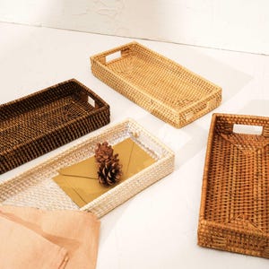 May include: Four rectangular woven rattan trays in varying shades of light tan, medium brown, dark brown, and white. Each tray features integrated side handles and a structured base. One white tray contains a brown envelope and two pinecones.