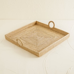 May include: A square woven tray with two round handles. The tray is made of natural woven rattan and has a light brown colour.
