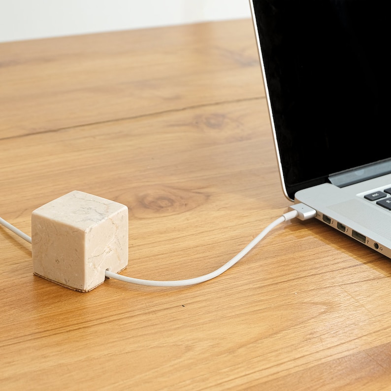 May include: A white marble cable organizer with a white cable running from it to a laptop on a wooden surface.