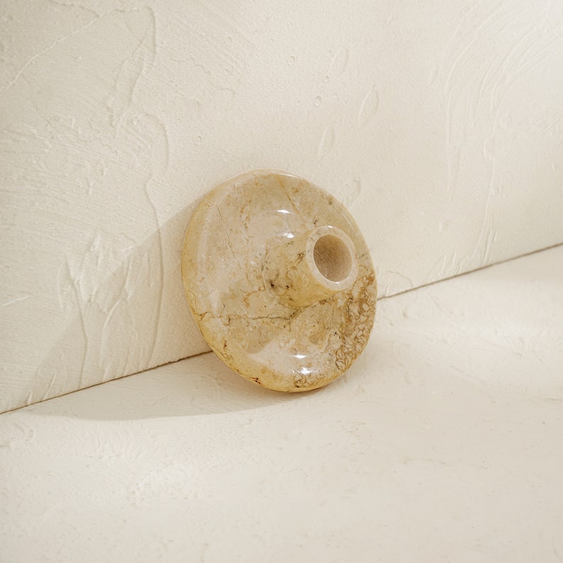 May include: A beige marble candle holder with a round base and a cylindrical opening for a candle.