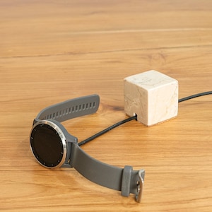 May include: A gray smartwatch with a black band is charging on a white marble charging stand. The charging cable is black and is routed through a hole in the stand.