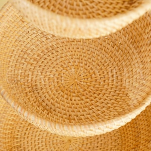 May include: A set of three woven baskets made from natural materials. The baskets are stacked on top of each other, with the largest basket at the bottom and the smallest basket at the top. The baskets are a light brown colour and have a circular shape.