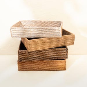 Pode incluir: Uma pilha de quatro bandejas retangulares trançadas com design de topo aberto. A bandeja superior é de cor bege claro, enquanto as três bandejas abaixo apresentam tons variados de marrom médio a escuro, com um padrão de trança consistente.
