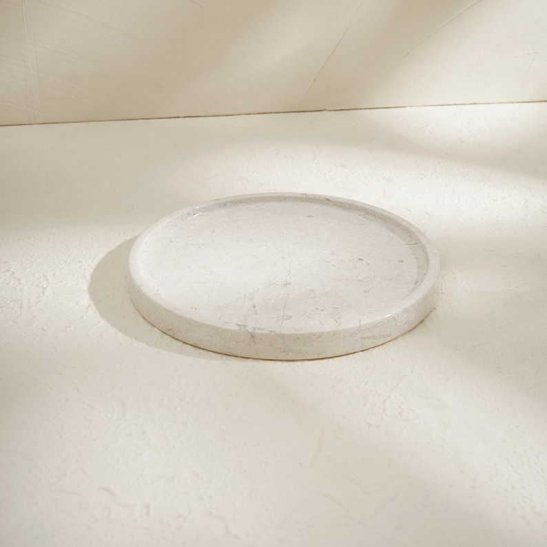 May include: A circular tray crafted from light beige stone with subtle grey veining. The tray features a smooth, polished surface and a raised rim, presenting a clean, flat design with a natural, mottled texture throughout the material.