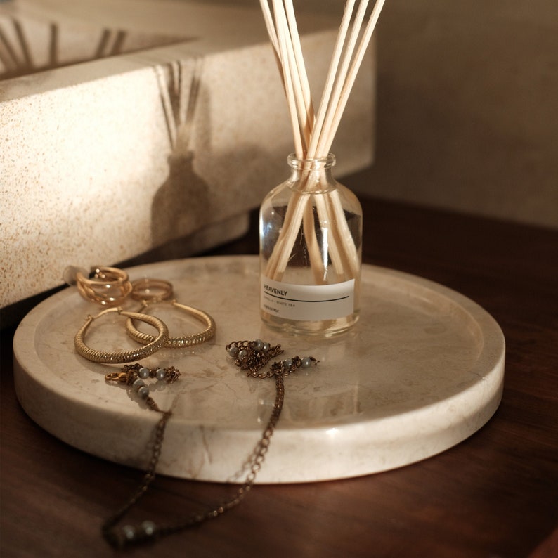 May include: A round beige marble tray holding a clear glass reed diffuser bottle with the label HEAVENLY, several light-colored wooden reeds, a pair of gold-toned textured hoop earrings, and a delicate gold-toned chain necklace with small gray beads.