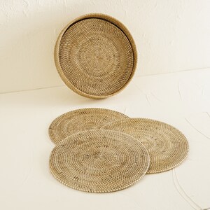 May include: Set of four round woven rattan placemats and a matching larger woven rattan tray. The placemats and tray are a light brown color.