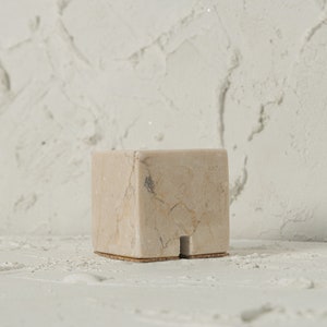 May include: A small, square, beige marble pedestal with a cork base. The pedestal has a natural, veined pattern and is perfect for displaying small objects.