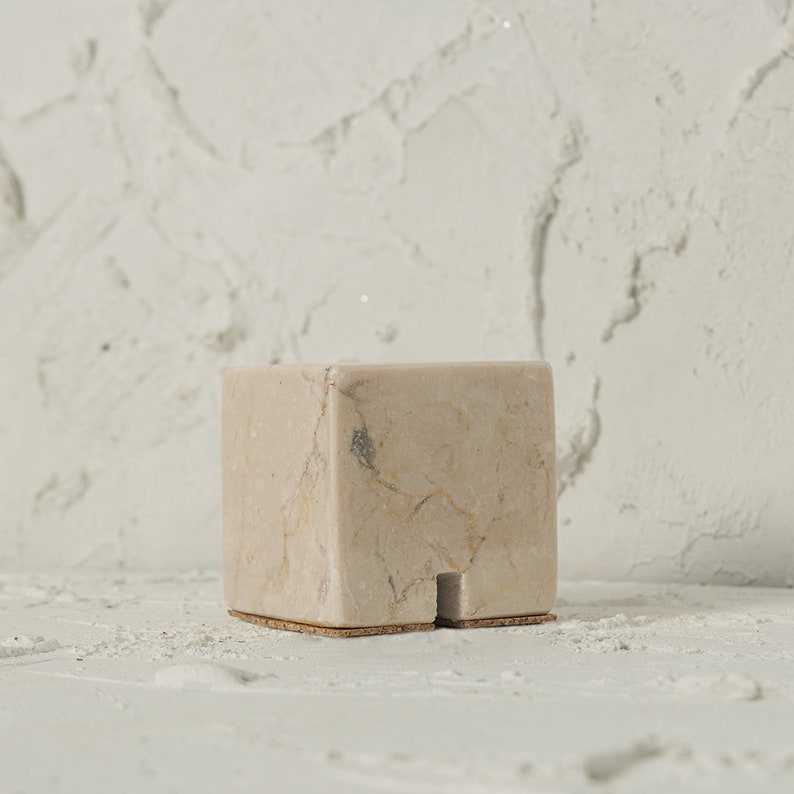 May include: A small, square, beige marble pedestal with a cork base. The pedestal has a natural, veined pattern and is perfect for displaying small objects.