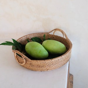 Op de afbeelding: Een ronde geweven rieten dienblad met twee groene mango's erin. Het dienblad heeft twee handvatten aan de zijkanten.