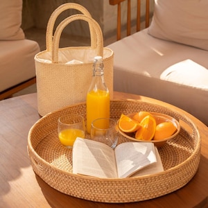 May include: A round woven rattan tray containing an open book, a glass bottle filled with orange juice, a small glass of orange juice, and a ceramic bowl holding several orange slices, positioned next to a tall woven straw tote bag.