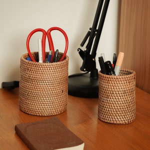 May include: Two woven wicker pencil holders, one filled with red scissors, pencils, and pens, and the other with black and grey pens and a pink highlighter. The holders are on a wooden surface.