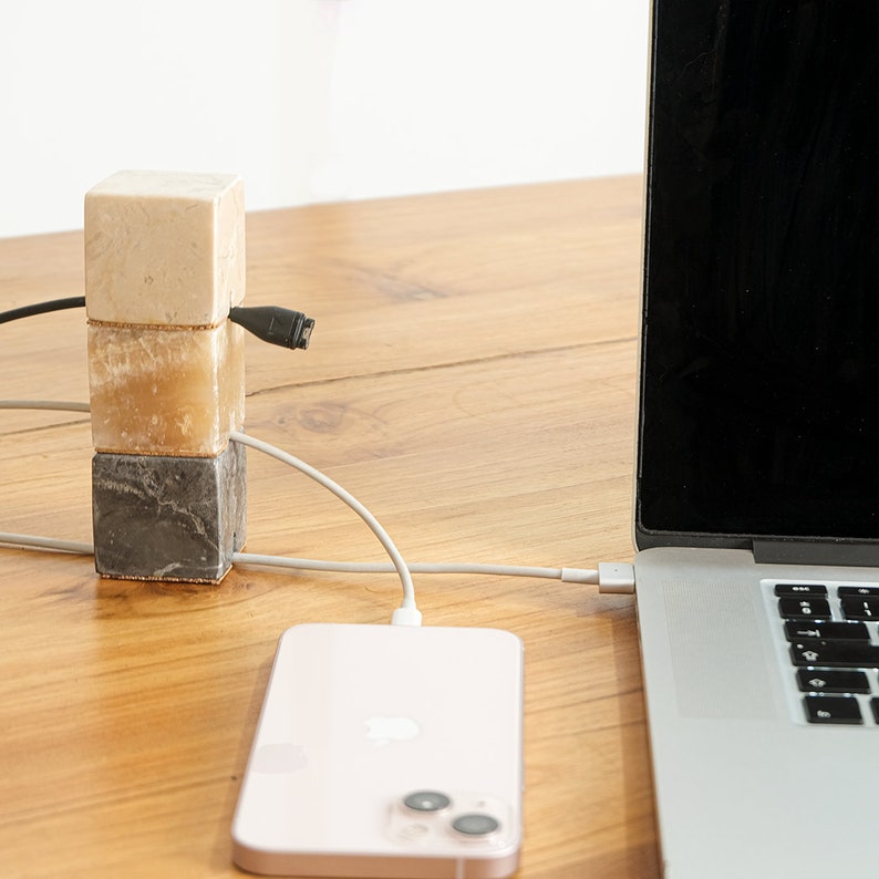 May include: A white, beige, and gray marble cable organizer with three cords running through it. A pink iPhone is charging on a wooden table next to a silver laptop.
