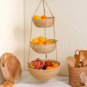 May include: A three-tiered hanging fruit basket made of woven natural material, with a variety of fruits including oranges, apples, dragon fruit, and a red apple. The basket is hanging from a white wall and is in a kitchen setting.