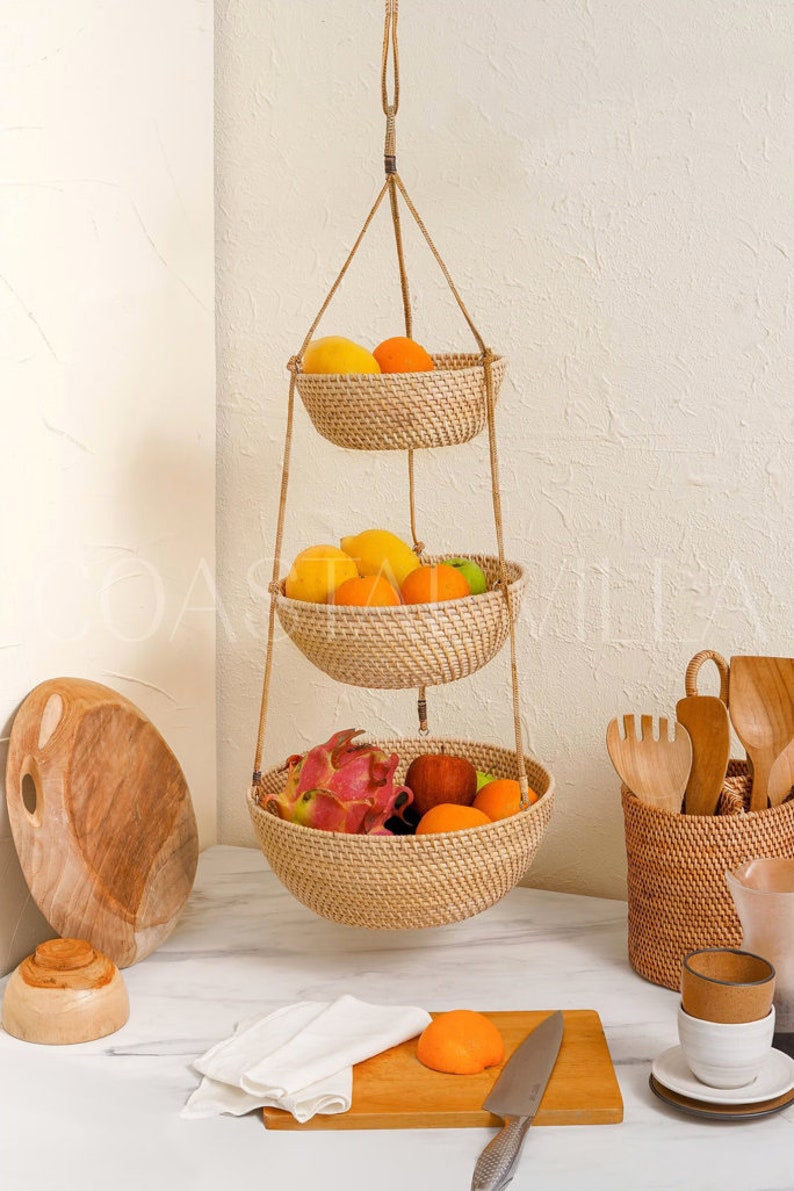 May include: A three-tiered hanging fruit basket made of woven natural material, with a variety of fruits including oranges, apples, dragon fruit, and a red apple. The basket is hanging from a white wall and is in a kitchen setting.