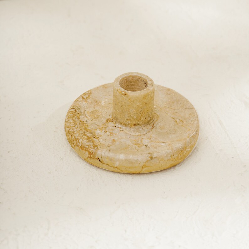 May include: A beige marble candle holder with a round base and a cylindrical top. The holder has a natural, veined texture.