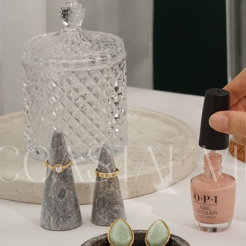May include: Two gray marble ring holders with gold rings on top. A bottle of O.P.I. nail lacquer in a light pink color is on a white marble surface. Two green stone earrings are in a small bowl.