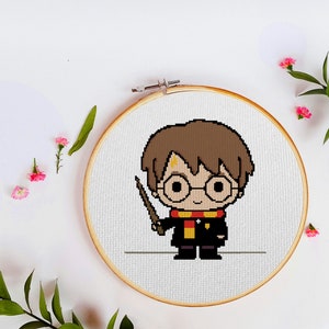 May include: A white embroidery hoop with a cross-stitch design of a cartoon Harry Potter character holding a wand. The character is wearing a black robe with a red and yellow scarf.