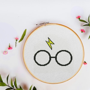 May include: A white embroidery hoop with a yellow lightning bolt and black glasses cross-stitched on it.