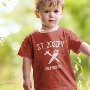 May include: A young boy wearing a red t-shirt with the text "ST. JOSEPH ORA PRO NOBIS" and a graphic of a hammer and saw. He is standing on a path in a grassy area.