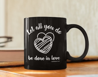 Könnte beinhalten: Schwarze Keramik-Kaffeetasse mit einem weißen Herzen und dem Text "Let all you do be done in love 1 Corinthians 16:14"