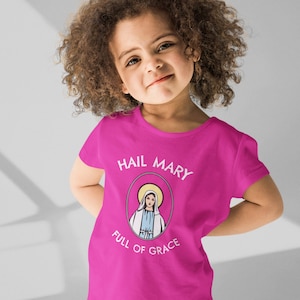 May include: A pink t-shirt with a white graphic of the Virgin Mary. The graphic says "Hail Mary Full of Grace".
