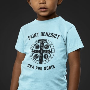 May include: A light blue t-shirt with a black print of Saint Benedict's Medal and the words "Saint Benedict" and "Ora Pro Nobis".