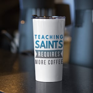 Puede incluir: Taza de viaje blanca con un dise&ntilde;o azul y gris que dice "Ense&ntilde;ar a los santos requiere m&aacute;s caf&eacute;".