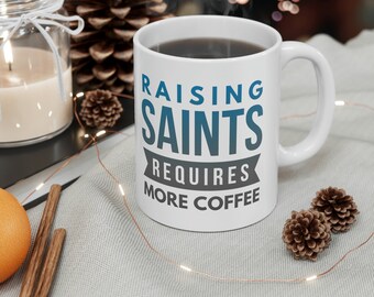 May include: White ceramic coffee mug with the text "Raising Saints Requires More Coffee" in blue and grey lettering. The mug is filled with coffee and steam is rising from the top.