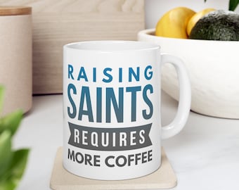 May include: White ceramic coffee mug with the text "Raising Saints Requires More Coffee" in blue and grey lettering.