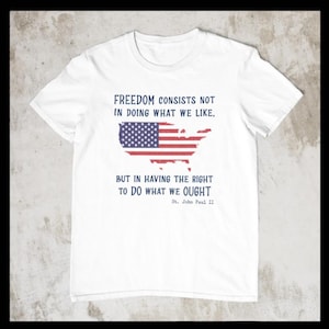 May include: White t-shirt with a distressed American flag design and a quote by St. John Paul II: "Freedom consists not in doing what we like, but in having the right to do what we ought."