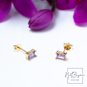 May include: A pair of gold earrings with small, rectangular, light purple gemstones. The earrings have a simple, minimalist design.