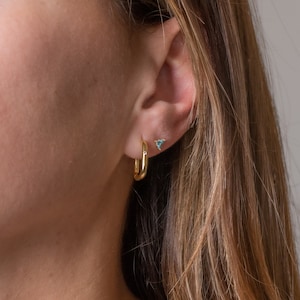 May include: A close-up of a person's ear with a gold hoop earring and a small gold triangle earring with turquoise stones.