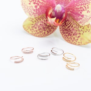 Puede incluir: Una colección de seis anillos en forma de espiral en tonos oro rosa, plata y oro. Cada anillo presenta una pequeña barra curva engastada con diminutas piedras transparentes. Los anillos están dispuestos por pares con una orquídea de fondo.