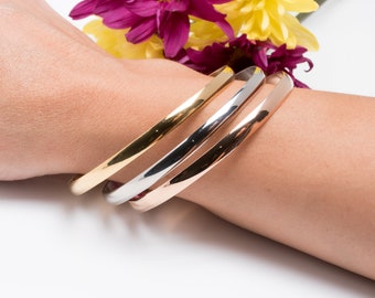 May include: Three gold, silver, and rose gold bangle bracelets stacked on a wrist.