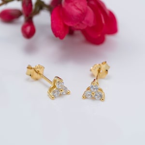 Könnte beinhalten: Ein Paar goldene Ohrstecker mit drei kleinen, klaren Zirkonia-Steinen, die in einer dreieckigen Form gefasst sind.