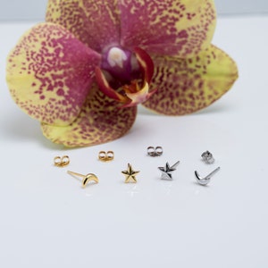 May include: Four pairs of gold and silver stud earrings. The earrings are shaped like stars and crescent moons. The earrings are displayed on a white surface with a pink and yellow orchid in the background.