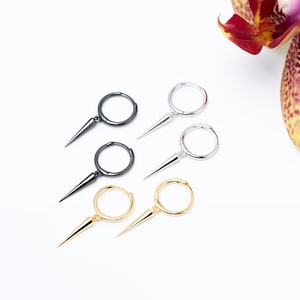 May include: An overhead shot showcases five hoop earrings with a spike pendant. The earrings are in three colors: black, silver, and gold. The background is white, with a partial view of a maroon and yellow orchid.