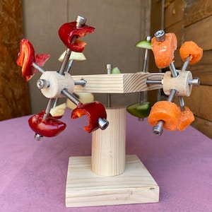 May include: A wooden stand with a rotating arm holding red and orange fruit slices. The stand is made of natural wood and has a simple design.