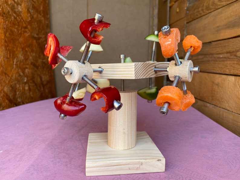 May include: A wooden stand with a rotating arm holding red and orange fruit slices. The stand is made of natural wood and has a simple design.