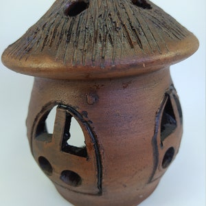 May include: A brown ceramic candle holder shaped like a house with a roof and cut-out windows. The candle holder has a rustic, handmade look.