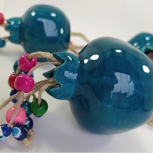 May include: Teal ceramic pomegranate ornaments with jute twine and colorful beaded accents. The pomegranates have a glossy finish and are adorned with pink, red, green, blue, and yellow beads. These decorative ornaments are ideal for home decor.