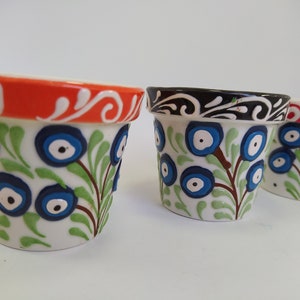 May include: Three small ceramic flower pots with blue evil eye designs and green leaves. The pots have different colored rims: orange, black, and red.