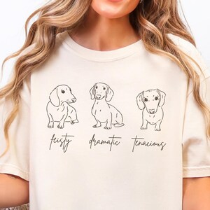 Può includere: T-shirt color crema con tre illustrazioni di bassotti in fila, ognuno in una posa diversa. Sotto i cani ci sono le parole "feisty, dramatic, tenacious" in un carattere scritto a mano. La maglietta è realizzata in materiale morbido.