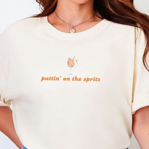 Op de afbeelding: Crèmekleurig T-shirt met de tekst "puttin' on the spritz" in oranje en een cocktailglas illustratie. Het shirt is gemaakt van zacht materiaal en heeft opgerolde mouwen.