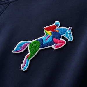May include: Navy blue sweatshirt with a colorful embroidered patch of a horse and jockey. The patch features a jumping horse with a rider, rendered in vibrant shades of blue, green, pink, and yellow. The sweatshirt has a classic crew neck design.