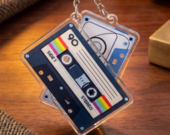 Puede incluir: Dos llaveros transparentes de acrílico con un diseño de cinta de cassette retro. Los llaveros presentan una cinta de cassette azul marino con rayas arcoíris, el número 90 y las palabras "SIDE 1" y "STEREO". Los llaveros están sujetos a cadenas plateadas.