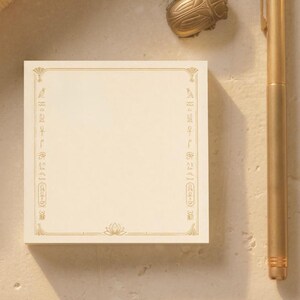 May include: A square cream-coloured notepad featuring a gold-toned border decorated with Egyptian hieroglyphs and a lotus flower at the bottom centre. A gold-toned metal pen and a small gold-toned beetle-shaped object are positioned nearby.